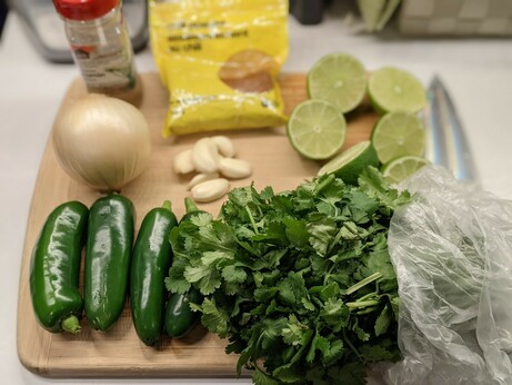 Cutting board holding jalapeños, a thick bunch of cilantro, six or so halved limes, a small pile of garlic cloves, a small sweet onion, cumin, and chili powder in a yellow No Name resealable bag.