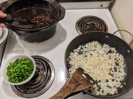Large frying pan with oil and diced onion in it.  To the left is the prep bowl with the diced jalapeños; behind is the black ceramic slow cooker insert full of beans and dark liquid.