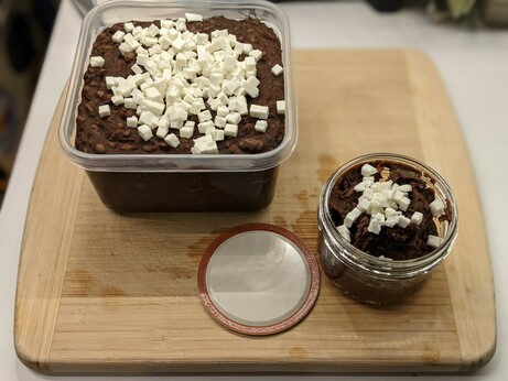 Large square plastic fridge dish full to the top with beans, plus a generous garnish of cubed white cheese on top.  In the foreground is a much smaller glass mason jar, garnished identically, holding what didn't fit in the larger tub.
