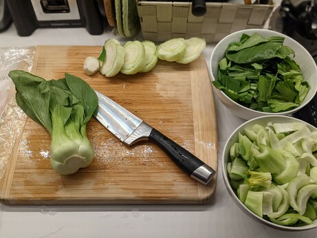Two bowls off to the right of the cutting board, one full of the large green leafy portions of the bok choy, generously sized, and one full of the white stem pieces cut into rather smaller pieces.  On the cutting board is a prep knife, the trimmed lowest stem bits of the vegetables, and one intact plant awaiting cleaning and chopping.
