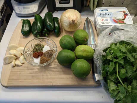 Mise en place on a bamboo cutting board: a pile of dark green jalapeño peppers, one medium sweet onion, five light green limes, a small pile of peeled garlic cloves, and a cut glass bowl with six small piles of seasonings in it (table salt, ground black pepper, cumin, monosodium glutamate, chili powder, and Mexican oregano).  To the right of the cutting board is a huge bunch of fresh cilantro; behind that is a small tub of feta cheese.